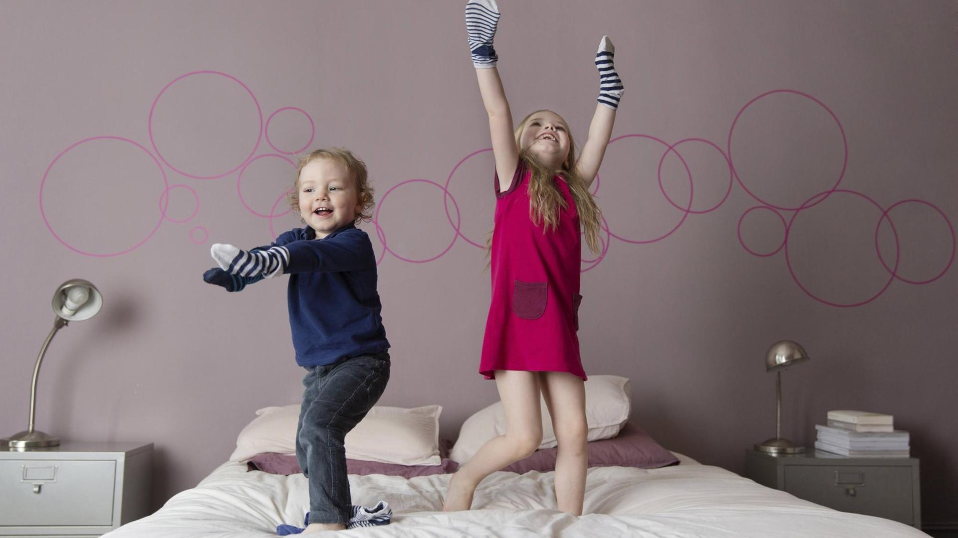Zwei Kinder stehen mit ihren Händen in Socken auf einem Bett.