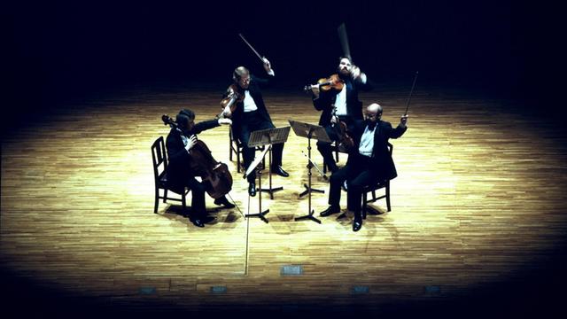 Vier Männer sitzen an ihren Instrumenten in einem schwarzen Raum, der eine Bühne sein könnte