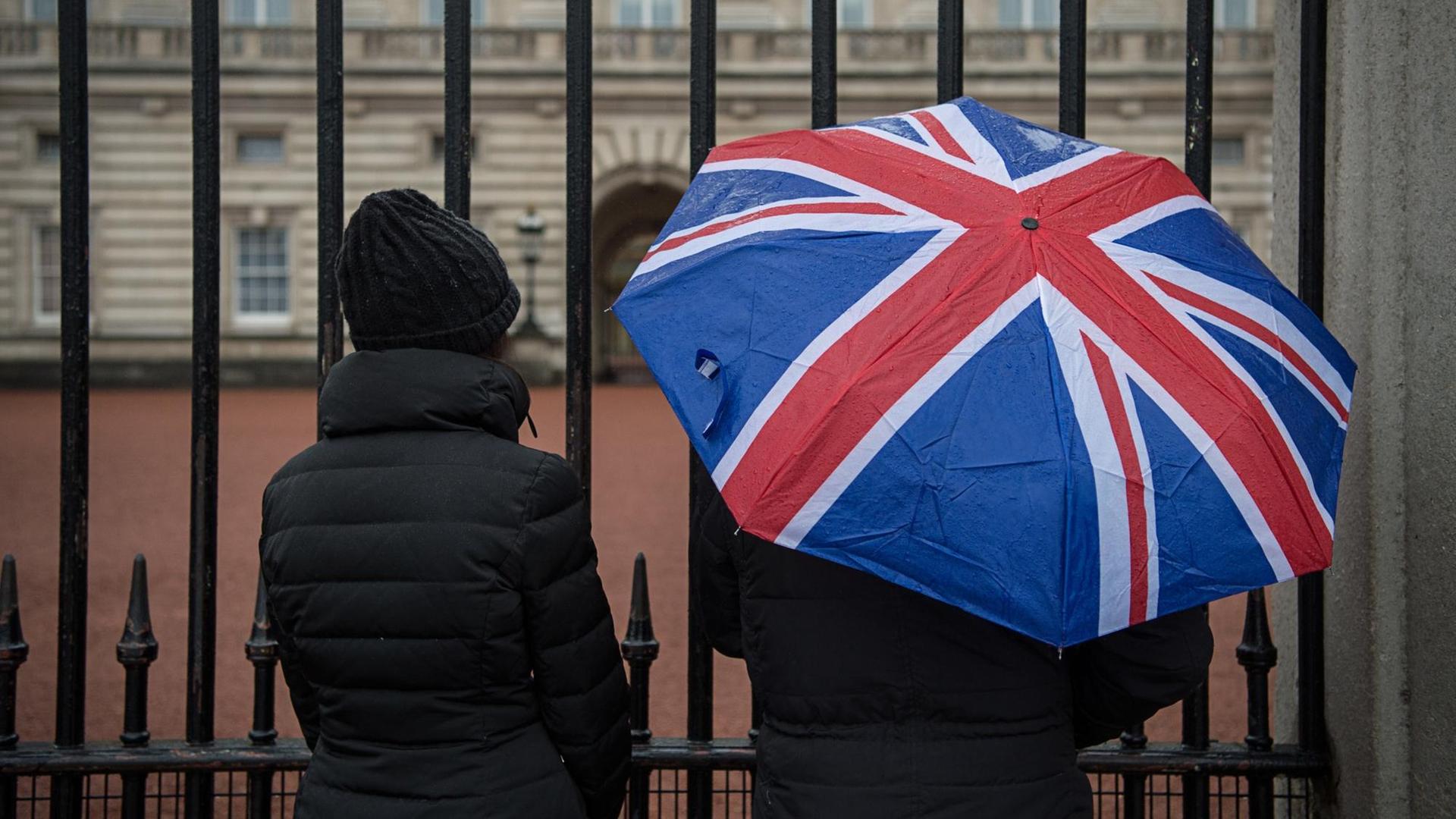 Touristen stehen mit einem Regenschirm in den Farben der Flagge des Vereinigten Königreich vor dem Buckingham Palace in London. Touristen stehen mit einem Regenschirm in den Farben der Flagge des Vereinigten Königreich vor dem Buckingham Palace in London.