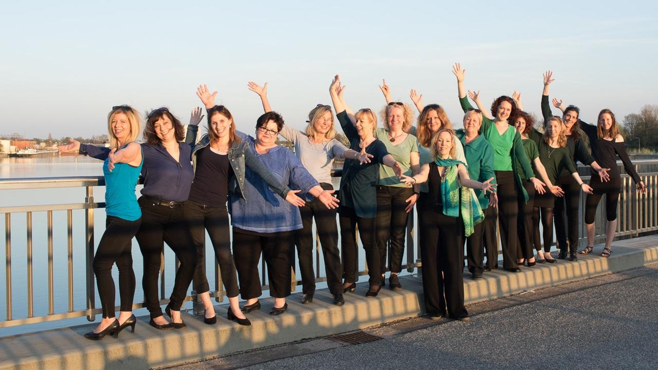 Die Frauen des Chores stehen aufgereiht am Geländer einer Brücke über einen Fluss. Die Frauen des Chores stehen aufgereiht am Geländer einer Brücke über einen Fluss.