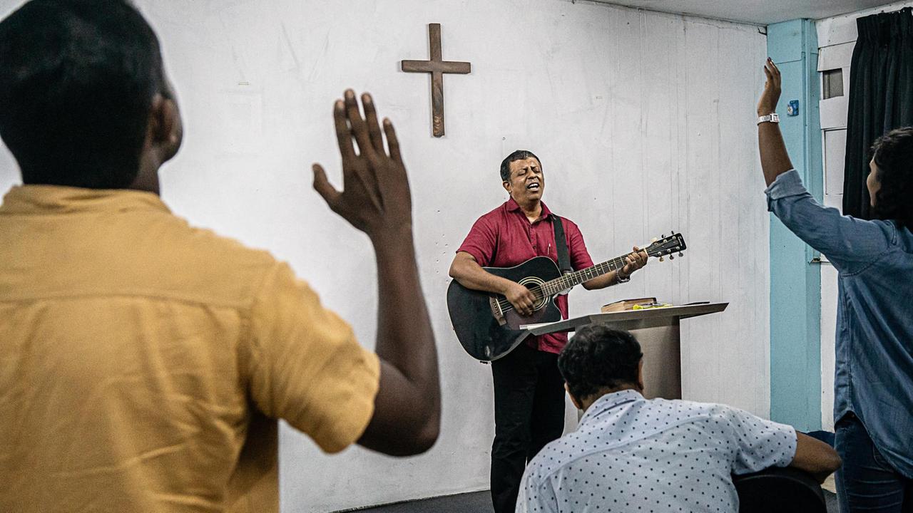 Tanuja Wahalatantri singt mit einer Gitarre um den Körper vor einer Wand, an der ein schlichtes Kreuz hängt. Um ihn herum stehen Menschen, die mit erhobenen Armen mitsingen.