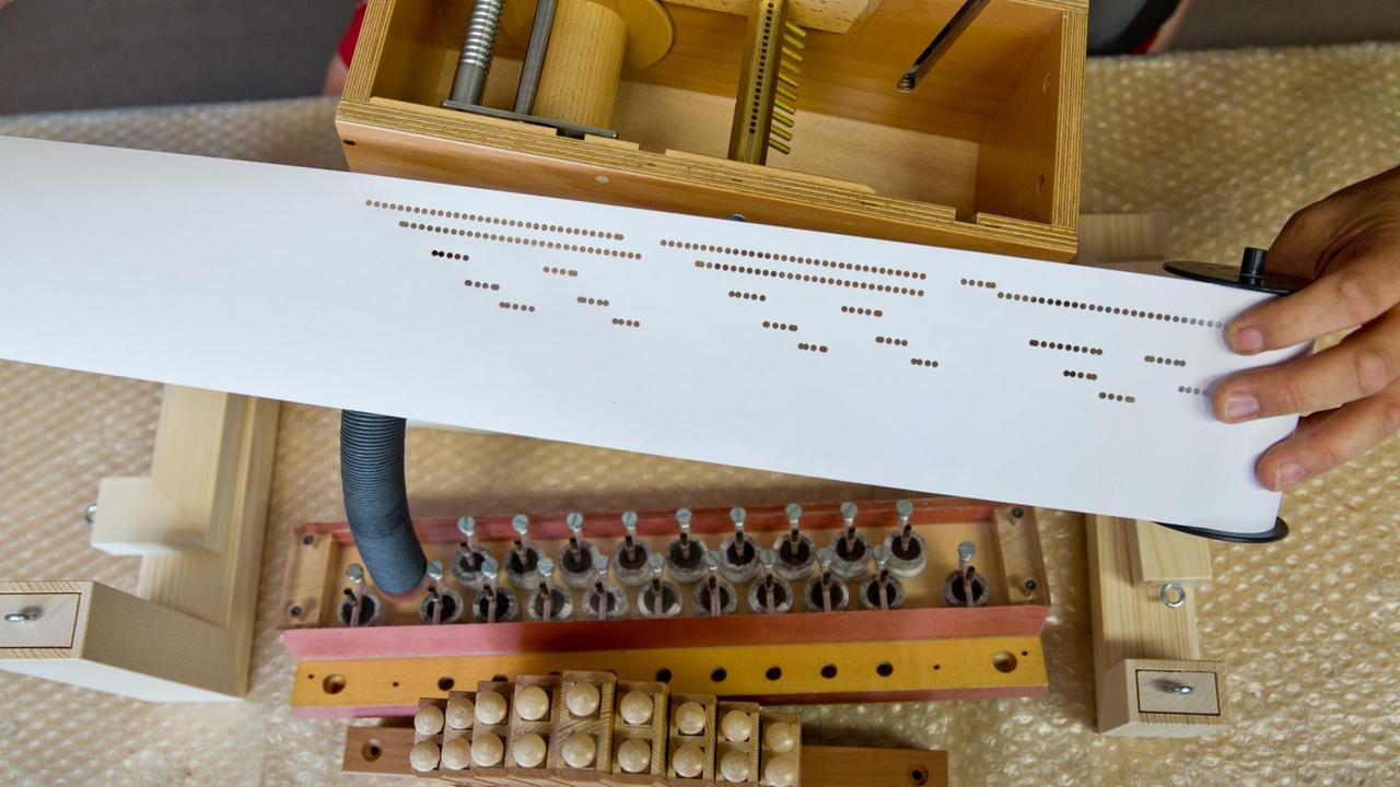 Das Notenband einer Drehorgel über einer in ihre Grundbauteile demontierten Drehorgel zu sehen. Das Notenband einer Drehorgel über einer in ihre Grundbauteile demontierten Drehorgel zu sehen.