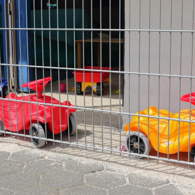 Zwei Bobbycars stehen hinter dem Zaun einer Kita.