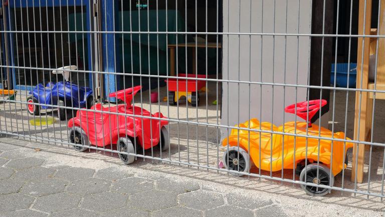 Zwei Bobbycars stehen hinter dem Zaun einer Kita.