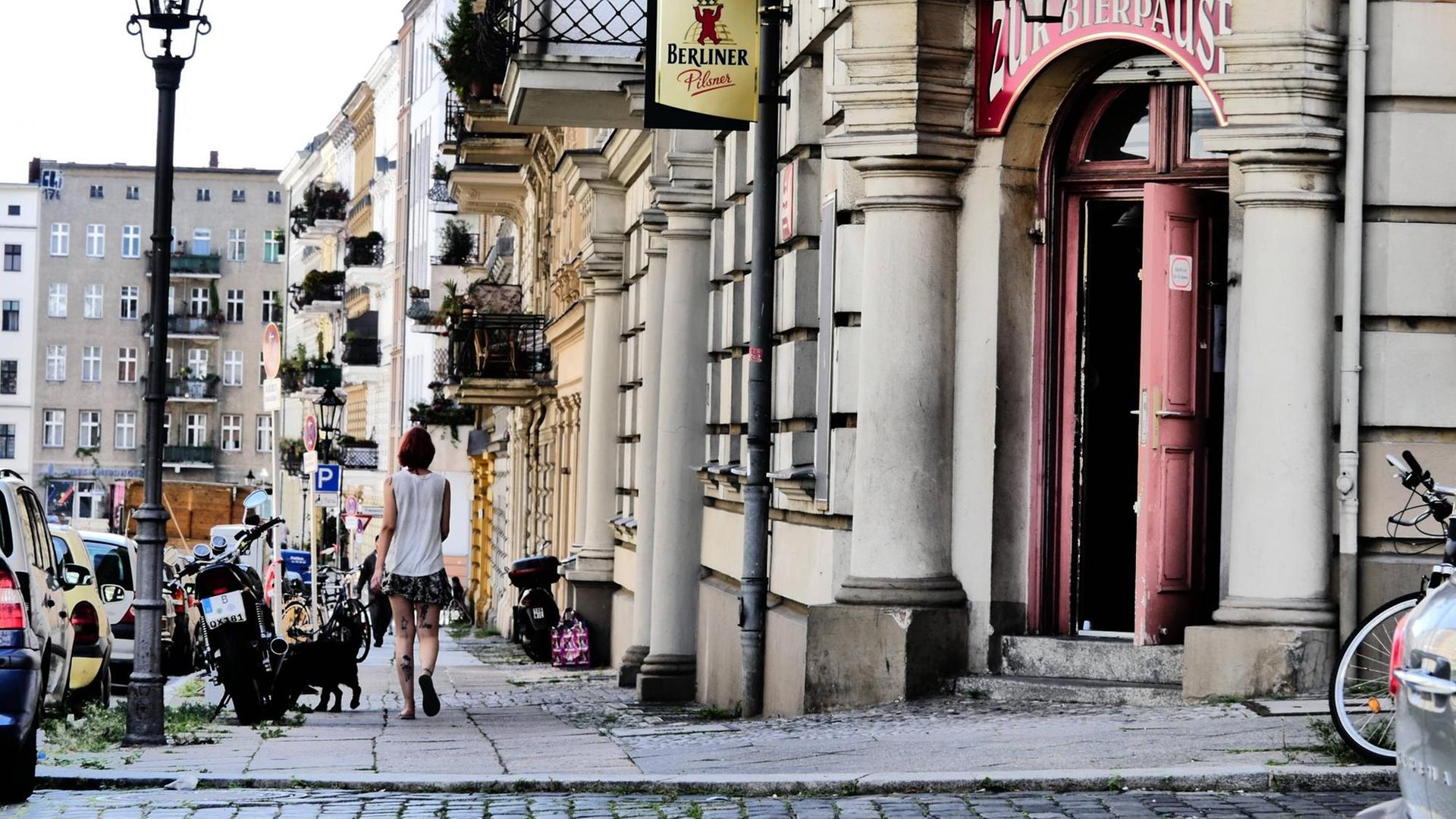 Eine Frau geht in Berlin Kreuzberg mit einem Hund auf dem Bürgersteig. Im Hintergund Häuser, rechts der Eingang zu einer Kneipe. Eine Frau geht in Berlin Kreuzberg mit einem Hund auf dem Bürgersteig. Im Hintergund Häuser, rechts der Eingang zu einer Kneipe.
