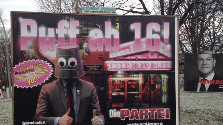 Großes Wahlplakat auf einer Grünfläche mit der Forderung: "Puff ab 16" und einem Bild von einem Mann, der einen Eimer auf dem Kopf trägt, im Hintergrund ist ein Bordell zu sehen.