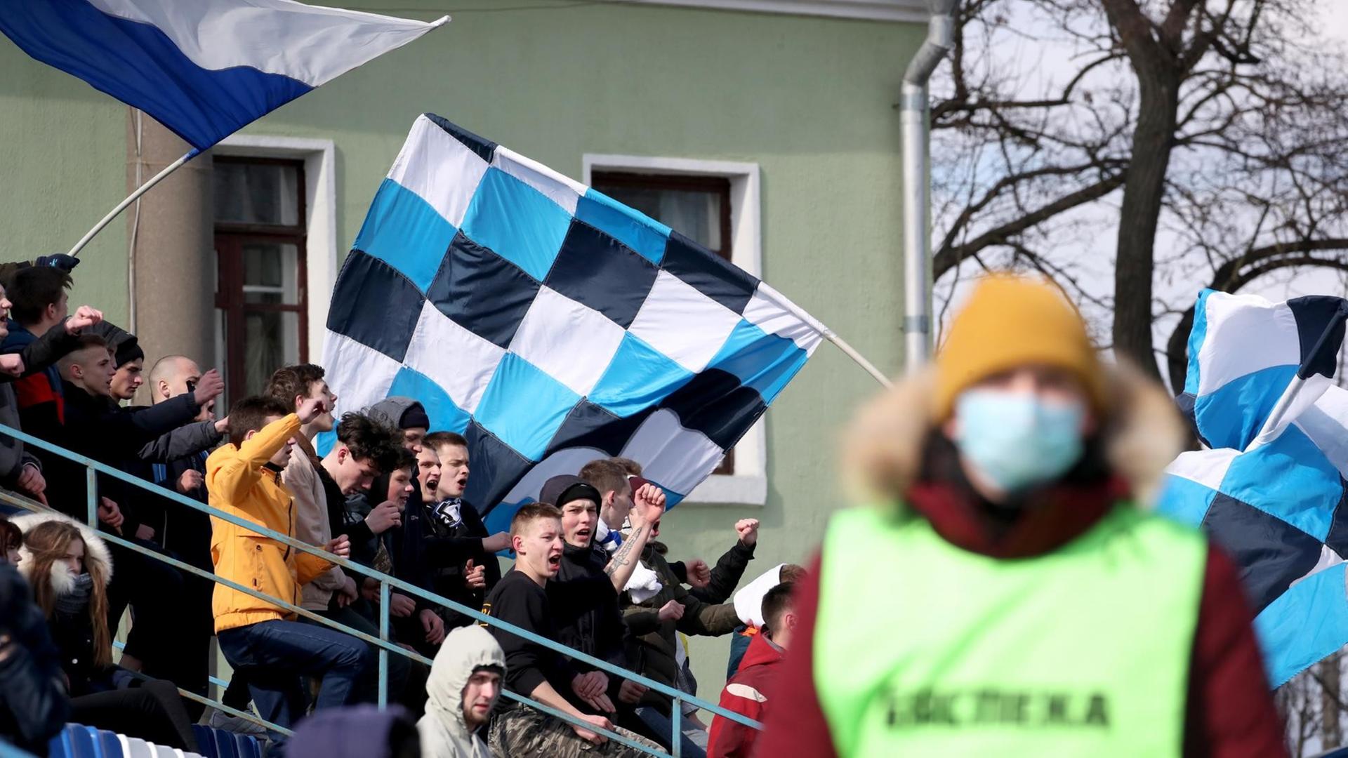 Fans im Fanblock beim Fußballspiel zwischen FC Slutsk und Slavia Mazyr, im Vordergrund ein Ordner mit einem Mundschutz Fans im Fanblock beim Fußballspiel zwischen FC Slutsk und Slavia Mazyr, im Vordergrund ein Ordner mit einem Mundschutz