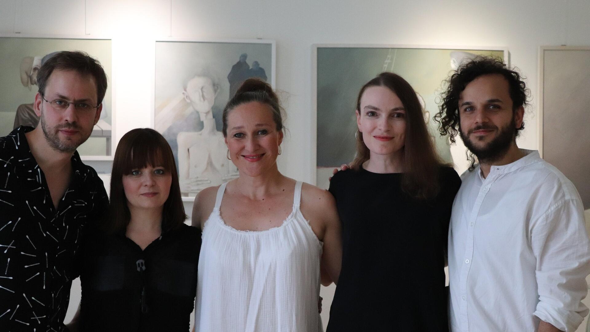 Fünf Perosnen stehen vor einer Wand. Dort hängen drei beleuchtet Kunstwerke Fünf Perosnen stehen vor einer Wand. Dort hängen drei beleuchtet Kunstwerke