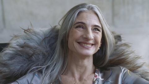 Ein Frau mit langen, glatten grauen Haaren sitzt lachend auf einem Sofa.
