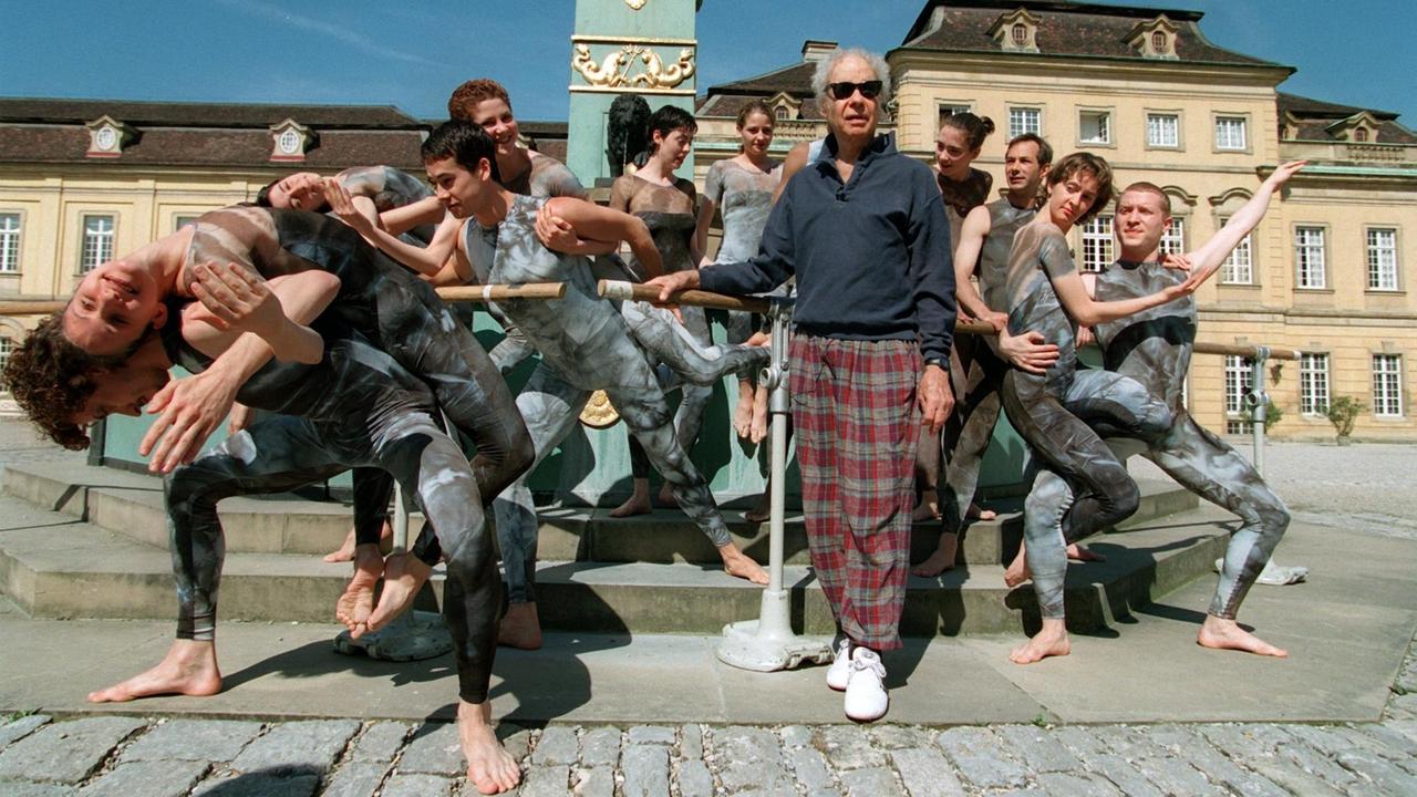 Merce Cunningham steht auf dem Platz vor dem Ludwigsburger Schloss, hinter und neben ihm sein Ensemble in dunklen, engen Kostümen in Tanzposen. Merce Cunningham steht auf dem Platz vor dem Ludwigsburger Schloss, hinter und neben ihm sein Ensemble in dunklen, engen Kostümen in Tanzposen.