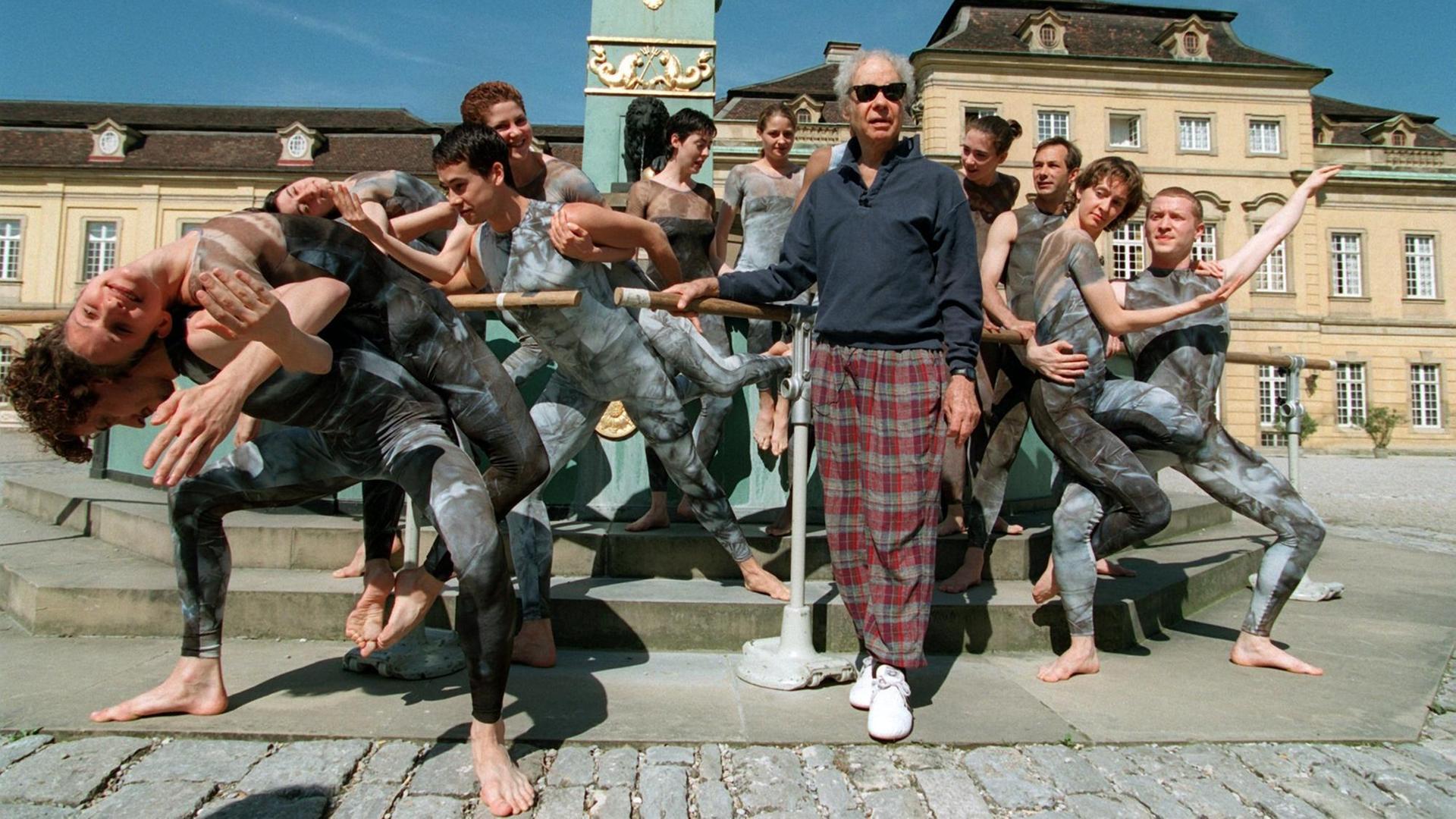 Merce Cunningham steht auf dem Platz vor dem Ludwigsburger Schloss, hinter und neben ihm sein Ensemble in dunklen, engen Kostümen in Tanzposen. Merce Cunningham steht auf dem Platz vor dem Ludwigsburger Schloss, hinter und neben ihm sein Ensemble in dunklen, engen Kostümen in Tanzposen.