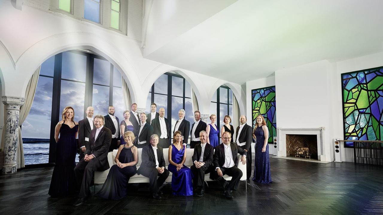 Das britische Ensemble "The Sixteen" präsentiert sich sitzend und stehend, in Konzertkleidung, in einer Halle, durch die Fenster im Hintergrund ist das Meer zu sehen