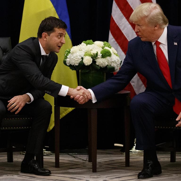 Donald Trump und Wolodymyr Selenskyj sitzen vor ihren Landesflaggen und geben sich die Hand.