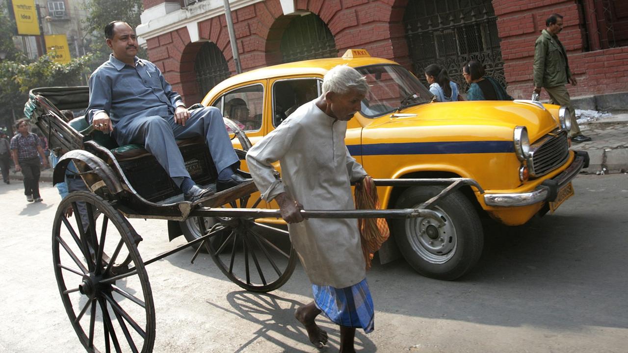 Ein älterer Rikscha-Fahrer zieht seine Rikscha mit einem Fahrgast durch eine Straße Kalkuttas, dahinter steht ein gelbes Taxi.