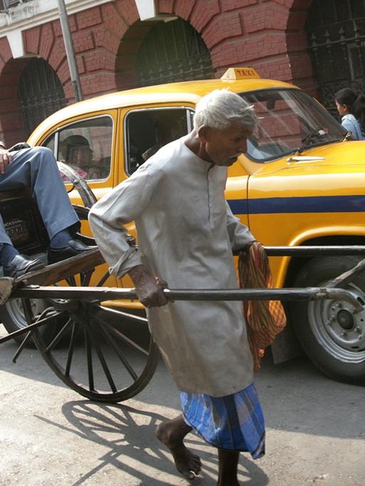 Ein alter Rikscha-Fahrer zieht seine Rikscha mit einem Fahrgast durch eine Straße Kalkuttas, dahinter steht ein gelbes Taxi.