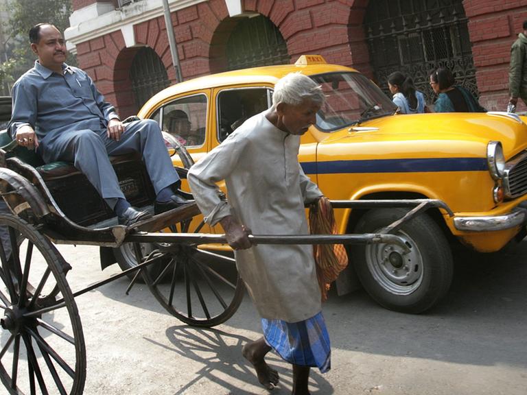 Ein älterer Rikscha-Fahrer zieht seine Rikscha mit einem Fahrgast durch eine Straße Kalkuttas, dahinter steht ein gelbes Taxi.