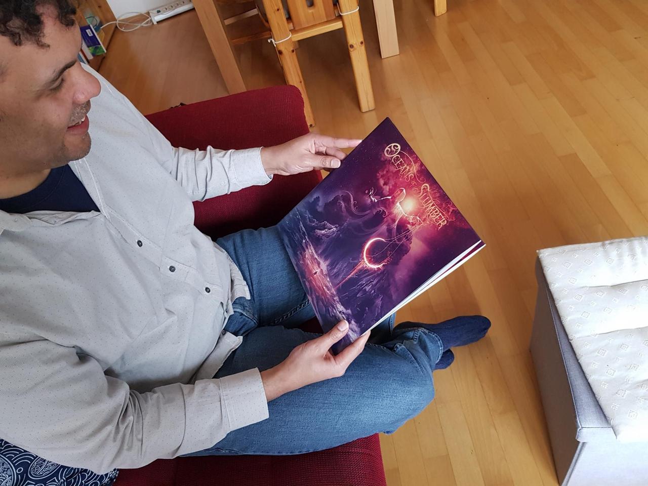 James A. Sullivan wurde von oben fotografiert und ist auf der linken Bildseite zu sehen. Er hält ein in rosa Farben gehaltenes, mystisch aussehendes Plattencover einer Heavy-Metal Band in den Händen. Das Cover nimmt den Mittelteil des Bildes ein. James A. Sullivan wurde von oben fotografiert und ist auf der linken Bildseite zu sehen. Er hält ein in rosa Farben gehaltenes, mystisch aussehendes Plattencover einer Heavy-Metal Band in den Händen. Das Cover nimmt den Mittelteil des Bildes ein.