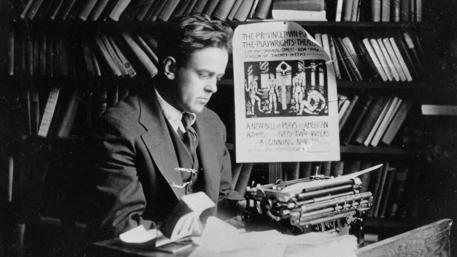 Ein schwarz-weißes Porträt-Foto zeigt den US-Journalisten John Silas Reed inmitten von Büchern Zeitungsstapeln und neben einer Schreibmaschine