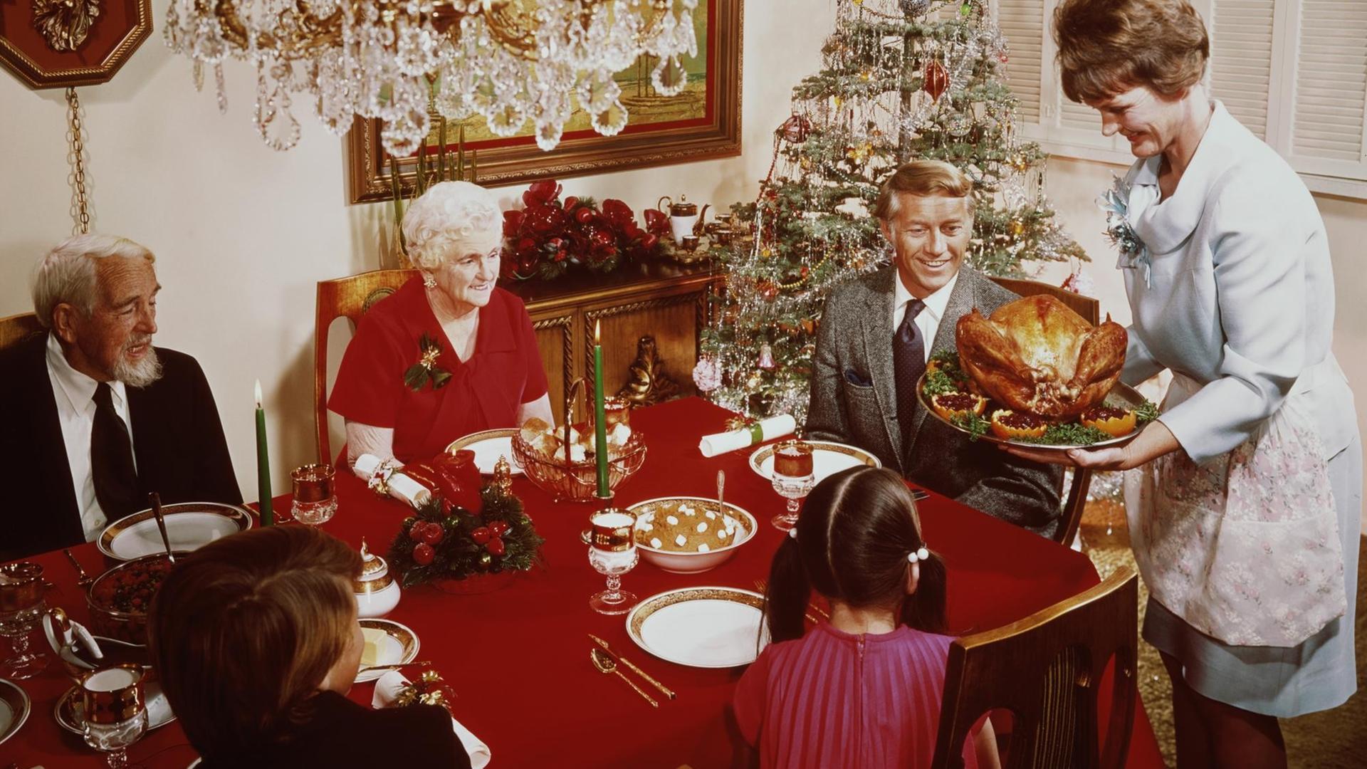Eine Aufnahe von 1965 zeigt eine Frau, die einen großen Truthahn zum Weihnachtsessen auf den Tisch bringt. Dort sitzen Großeltern und Kinder am gedeckten Tisch. Im Hintergrund ist ein geschmückter Weihnachtsbaum zu sehen.