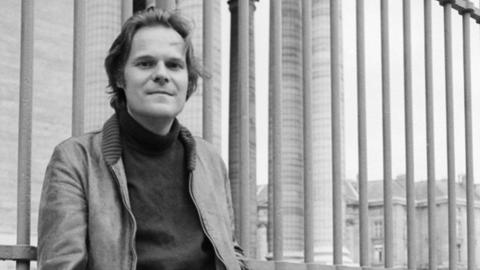 Schwarzweißfoto: Der Schriftsteller Peter Schneider lehnt lässig an einem Zaun.
