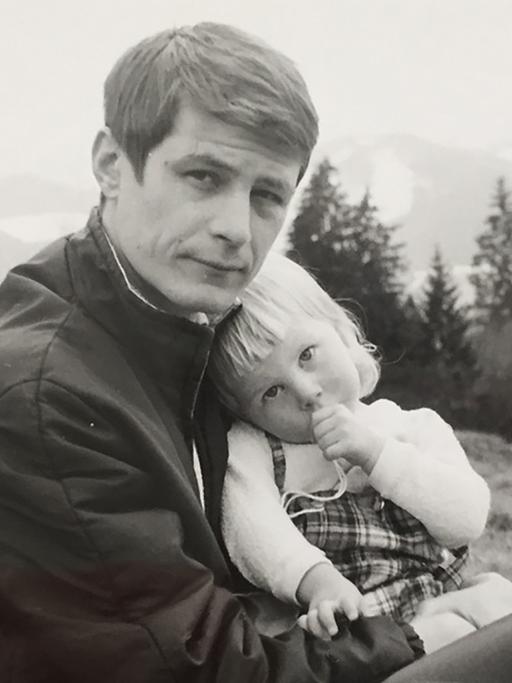 Vater und Tochter sitzen in den Bergen, die Tochter schmiegt sich an ihren Vater und nuckelt am Daumen.