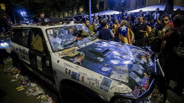 Ein Fahrzeug der spanischen Guardia Civil beschädigt steht nachts auf einer Straße in Barcelona. Daneben stehen Menschen, die für ein Unabhängigkeitsreferendum demonstrieren.