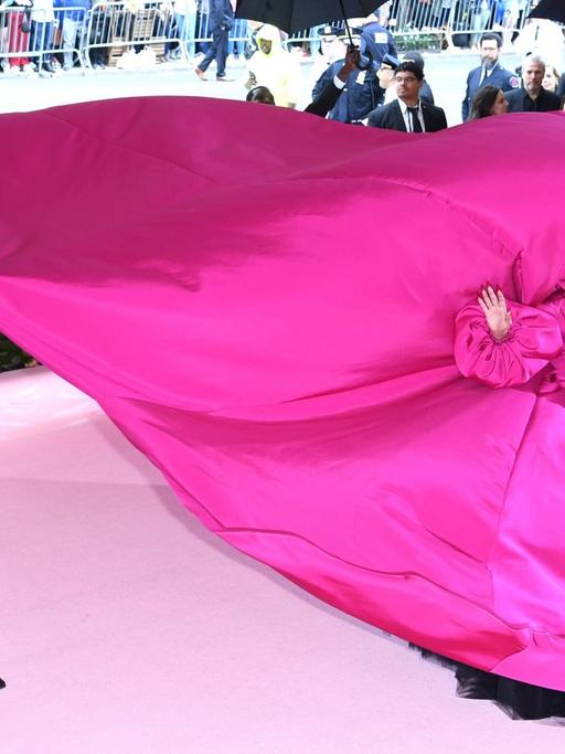 Lady Gaga auf der Met Gala in einem pink-violettem Kleid, dessen Schleppe von mehreren Männern getragen wird.
