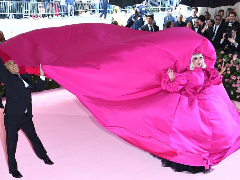Lady Gaga auf der Met Gala in einem pink-violettem Kleid, dessen Schleppe von mehreren Männern getragen wird.