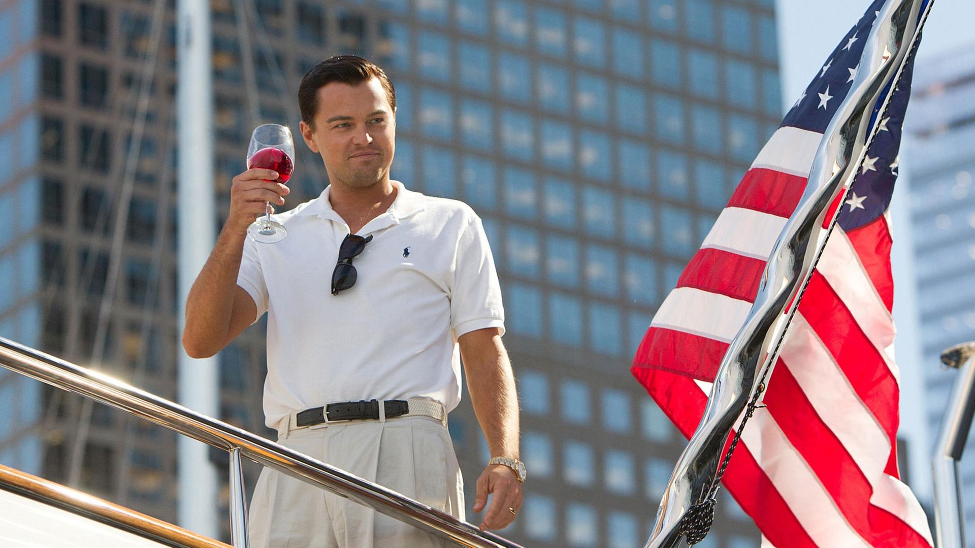 Leonardo DiCaprio steht neben einer US-Flagge auf einem Schiff und hält einen Drink hoch