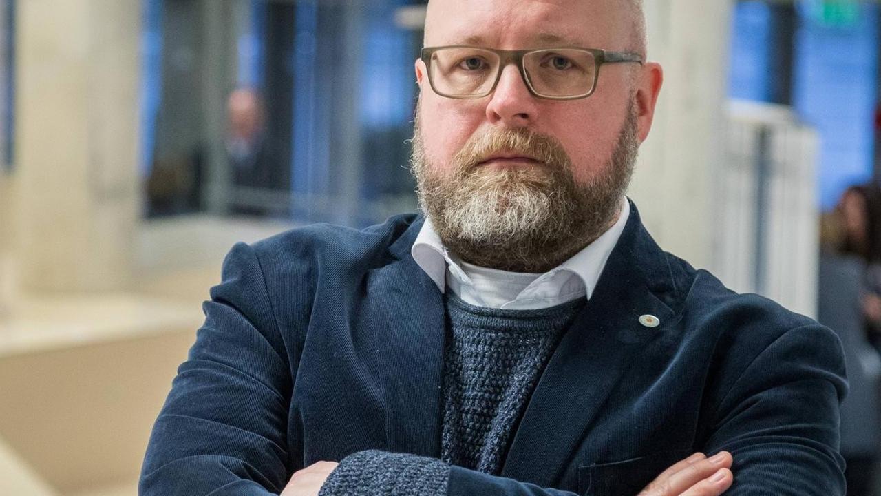 Ein Mann mit Vollbart und verschränkten Armen steht in einem Foyer und schau ernst in die Kamera. Ein Mann mit Vollbart und verschränkten Armen steht in einem Foyer und schau ernst in die Kamera.