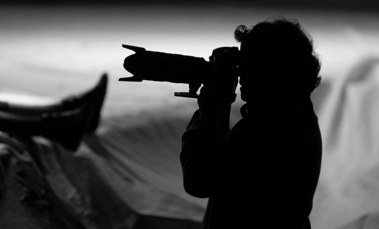 Im Gegenlicht ist eine Fotografin als Schattenriss zu erkennen, die eine Kamera mit einem langen Objektiv vor den Augen hat.