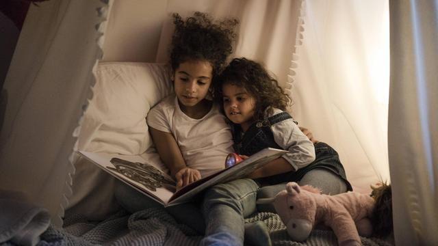Zwei kleine Mädchen, Schwestern, sitzen zusammen im Kinderzimmer unter einem Baldachin und blättern in einem Buch.