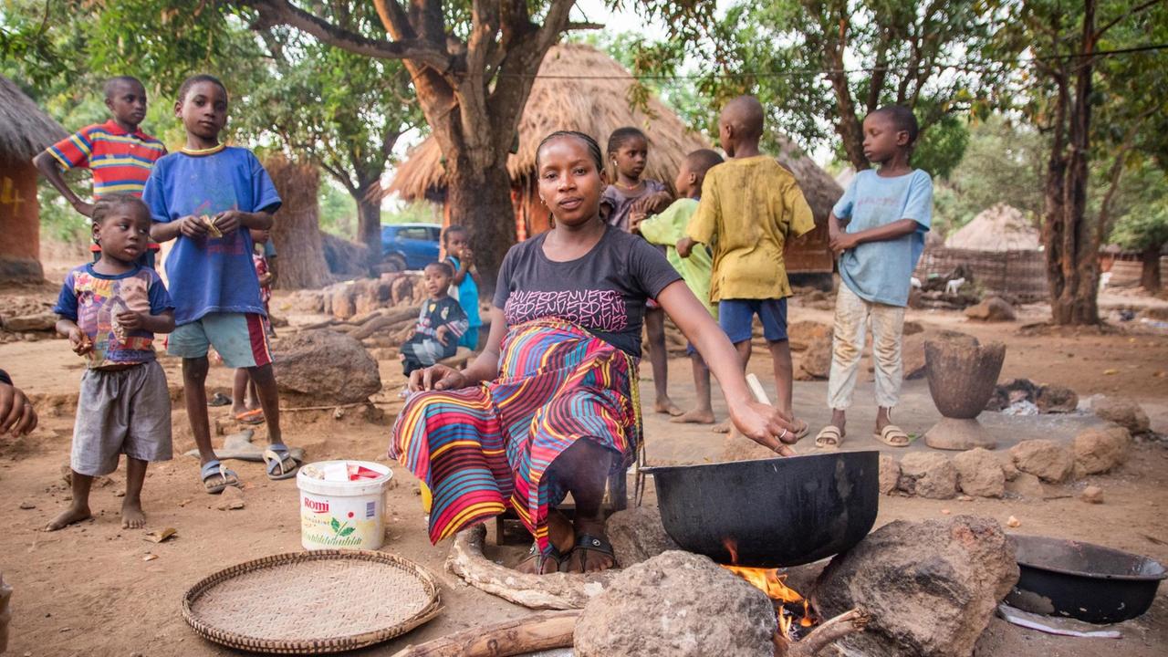 Dorfleben in Hamdallaye. Eine schwangere Frau sitzt an einer Kochstelle. Im Hintergrund ein afrikanisches Dorf und viele Kinder, die neugierig in die Kamera schauen. 