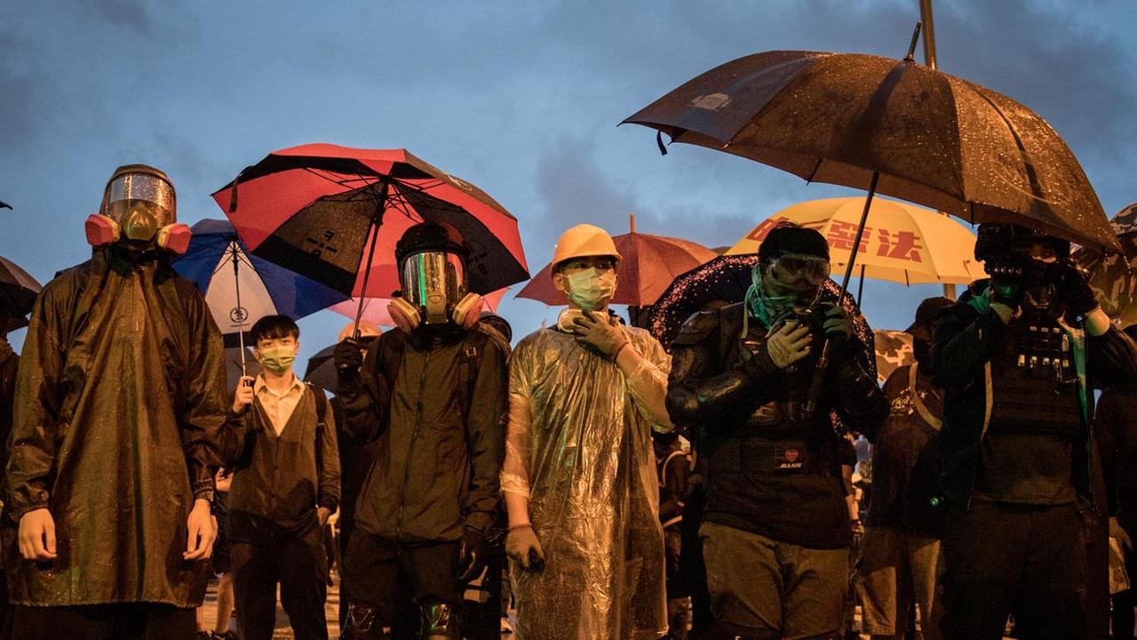 Demonstranten stehen im Regen, mit Regenschirmen und Schutzmasken vor dem Gebäude des Legislativrates, der Polizei gegenüber. 
