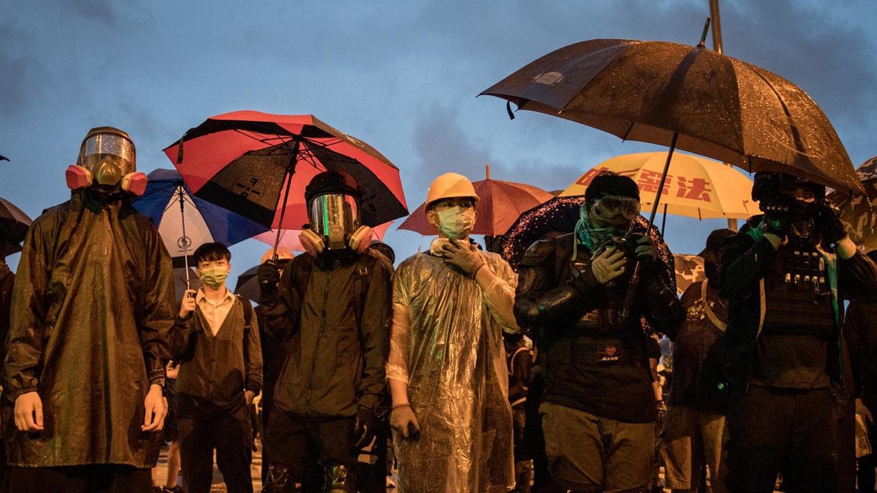 Demonstranten stehen im Regen, mit Regenschirmen und Schutzmasken vor dem Gebäude des Legislativrates, der Polizei gegenüber. Demonstranten stehen im Regen, mit Regenschirmen und Schutzmasken vor dem Gebäude des Legislativrates, der Polizei gegenüber.