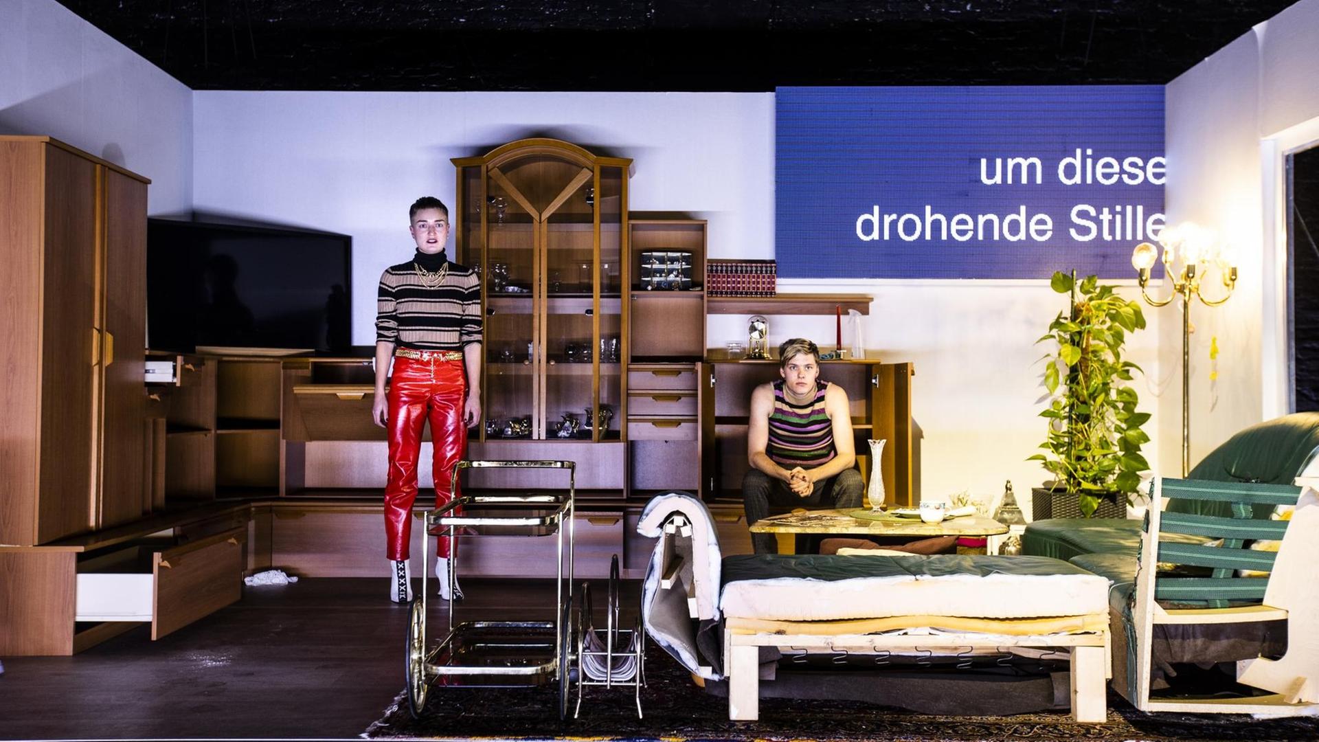 Szene aus dem Theaterstück "Tragödienbastard". Zu sehen ist eine Art Wohnzimmer mit zwei Menschen. Auf einer Art Bildschirm steht "um diese drohende Stille". Szene aus dem Theaterstück "Tragödienbastard". Zu sehen ist eine Art Wohnzimmer mit zwei Menschen. Auf einer Art Bildschirm steht "um diese drohende Stille".