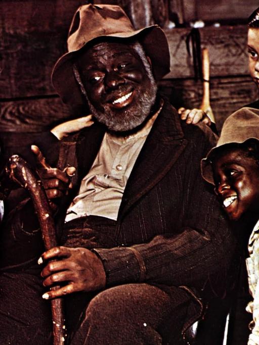 James Baskett als Onkel Remus sitz in einem Stuhl, umkreist von drei Kindern; Luana Patten als Ginny Favers, Bobby Driscoll als Johnny und Glenn Leedy als Toby.