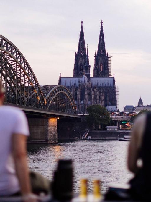Kölner Skyline mit Kölner Dom, im Vordergrund trinken zwei Menschen Bier.