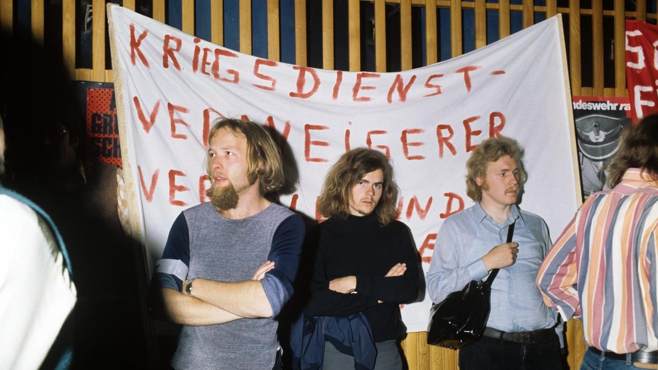 Die Aufnahme zeigt eine Protestkundgebung gegen den Wehrdienste von 1974: Drei junge Männer stehen vor einem Transparent auf dem "Kriegsdienstverweigerer" aufgemalt ist.