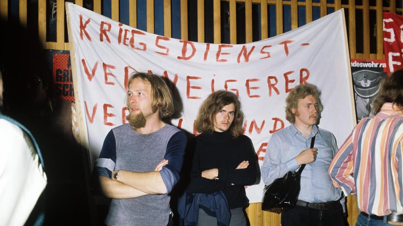 Die Aufnahme zeigt eine Protestkundgebung gegen den Wehrdienste von 1974: Drei junge Männer stehen vor einem Transparent auf dem "Kriegsdienstverweigerer" aufgemalt ist. Die Aufnahme zeigt eine Protestkundgebung gegen den Wehrdienste von 1974: Drei junge Männer stehen vor einem Transparent auf dem "Kriegsdienstverweigerer" aufgemalt ist.