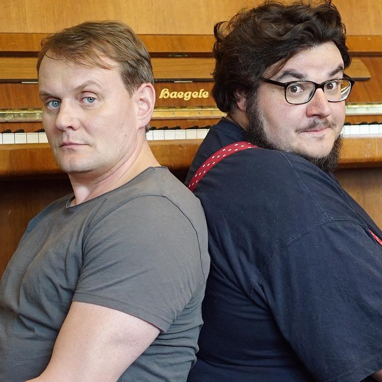 Devid Striesow und Axel Ranisch sitzen Rücken an Rücken vor einem Klavier.
