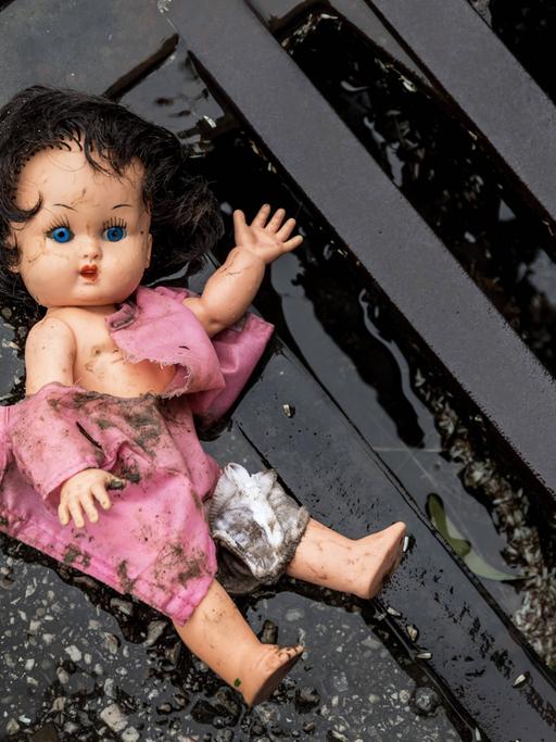 Eine Puppe liegt in dreckiger Bekleidung an einem Straßengully.