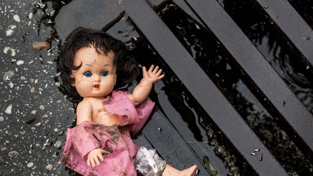 Eine Puppe liegt in dreckiger Bekleidung an einem Straßengully.