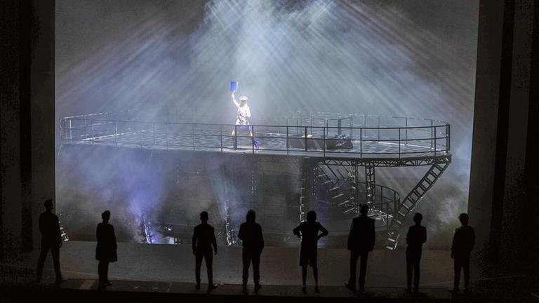 Auf einer Theaterbühne steht eine Frau im Matrosen-Look auf einem großen metallenen Schiffsrumpf und hält ein blaues Buch in die Höhe. Auf einer Theaterbühne steht eine Frau im Matrosen-Look auf einem großen metallenen Schiffsrumpf und hält ein blaues Buch in die Höhe.