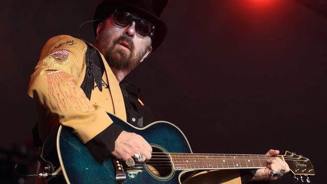 Der Musiker und Produzent Dave Stewart spielt mit Sonnenbrille und Hut im Rampenlicht auf einer Gitarre.