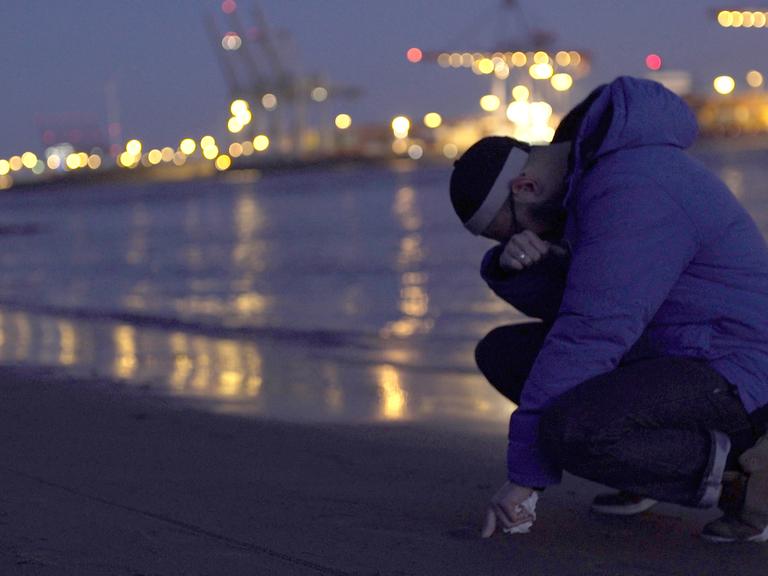 Der Protagonist Danisch hockt niedergeschlagen am Elbstrand. Im Hintergrund sind die Lichter vom Hafen zu erkennen.