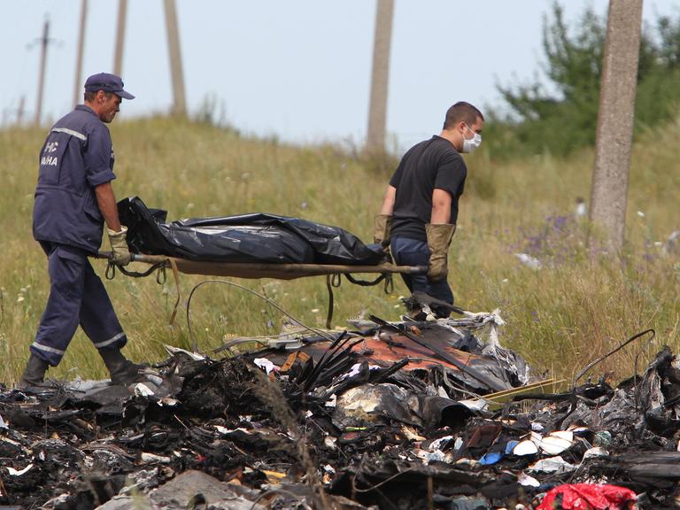 Hinter Trümmern tragen zwei ukrainische Arbeiter eine Bahre, auf der ein abgedecktes Todesopfer liegt.