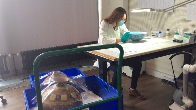 Katharina Steegmans sitzt an einem Arbeitstisch und restauriert eine blaue Schale. Über ihr und der Schale hängt eine Lampe und sie selbst trägt blaue Plastikhandschuhe, sowie einen weißen Kittel. Links neben ihr steht ein Rollwagen in dem sich bauchige, historische Vasen befinden Katharina Steegmans sitzt an einem Arbeitstisch und restauriert eine blaue Schale. Über ihr und der Schale hängt eine Lampe und sie selbst trägt blaue Plastikhandschuhe, sowie einen weißen Kittel. Links neben ihr steht ein Rollwagen in dem sich bauchige, historische Vasen befinden