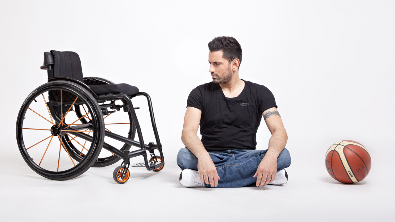 Tan Caglar sitzt im Schneidersitz auf dem Boden in einem hell ausgeleuchteten Fotostudio und schaut nach links auf einen Rollstuhl. Rechterhand liegt ein Baskettball.