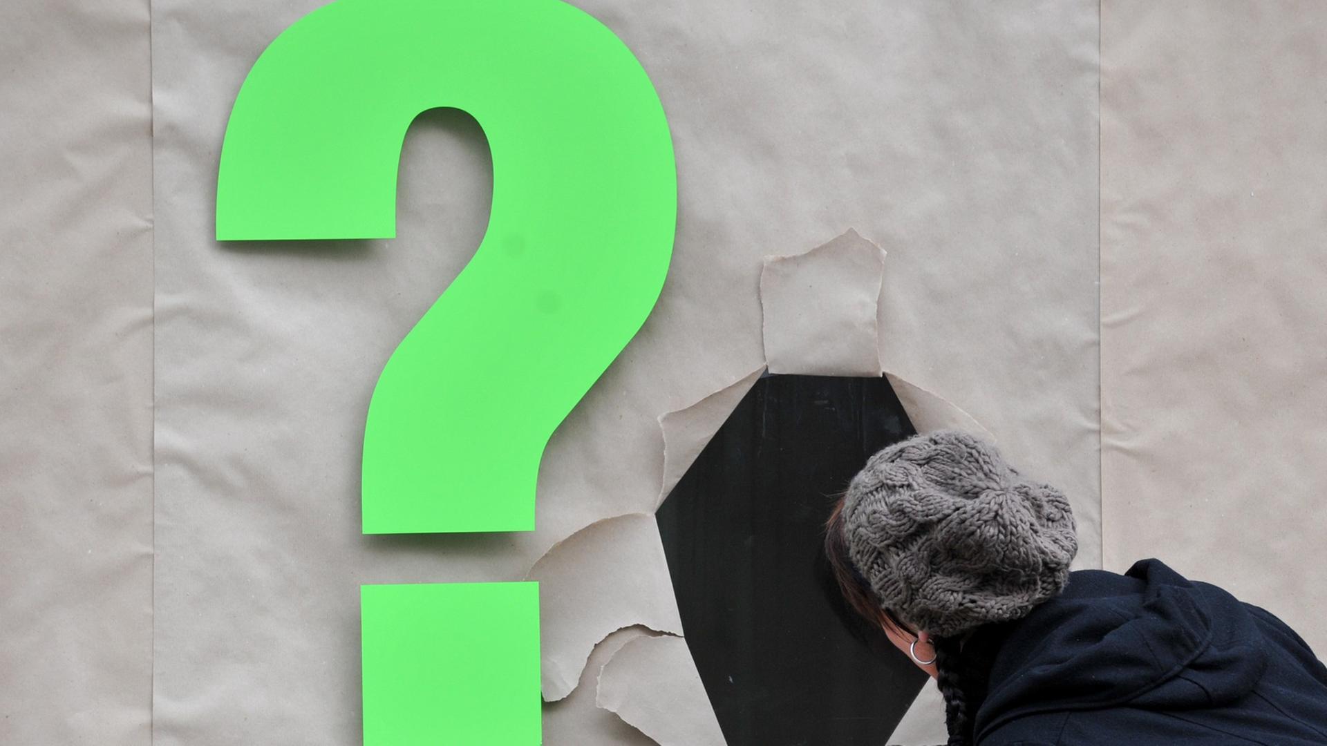 Eine Frau blickt an einem Fragezeichen vorbei durch eine aufgerissene Öffnung an einem mit Packpapier verdeckten Schaufenster. Eine Frau blickt an einem Fragezeichen vorbei durch eine aufgerissene Öffnung an einem mit Packpapier verdeckten Schaufenster.